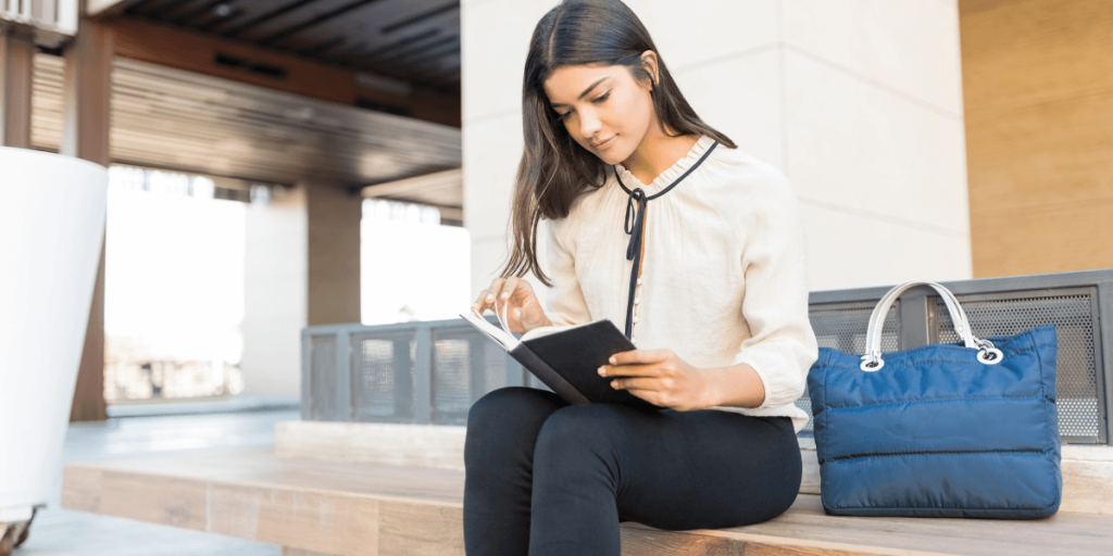 Young woman in a white blouse reads a book while seated on outdoor steps, a blue handbag beside her. The setting is modern and peaceful.
